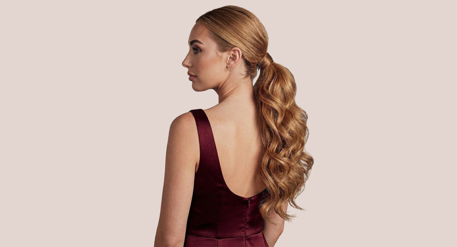 Une personne aux longs cheveux blonds ondulés, coiffés en queue de cheval basse, portant une robe bordeaux foncé sur fond neutre.