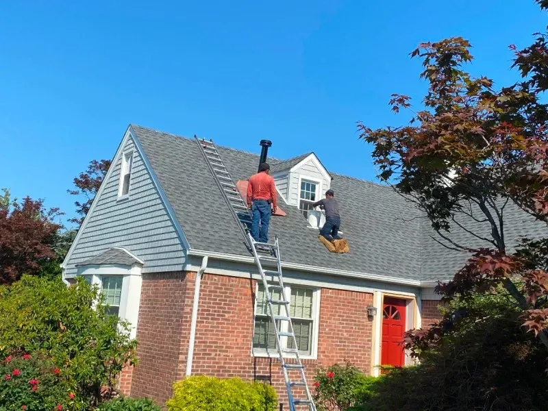 Two people are working on the grey shingled roof of a brick house with a red front door on a sunny day.