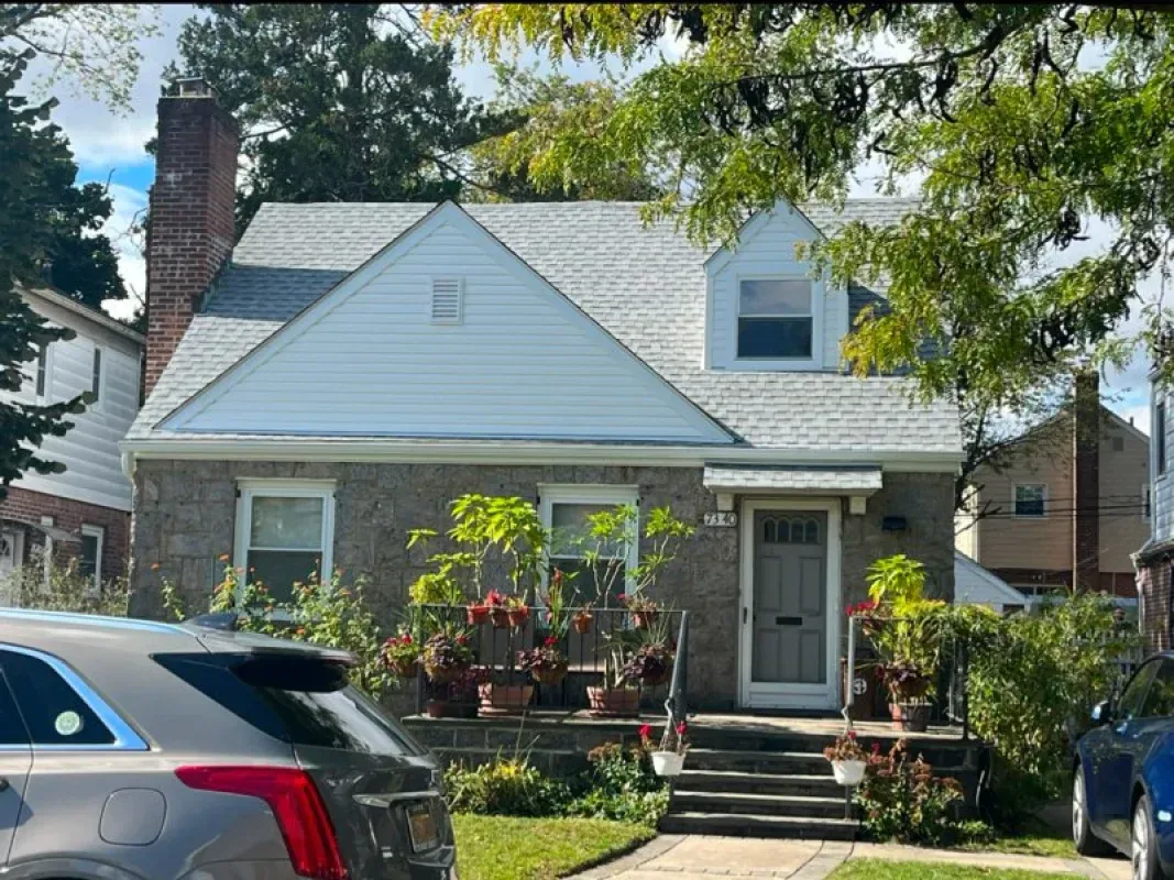 A stone house with white siding, a chimney, and a small front porch, surrounded by trees and plants in a residential area.