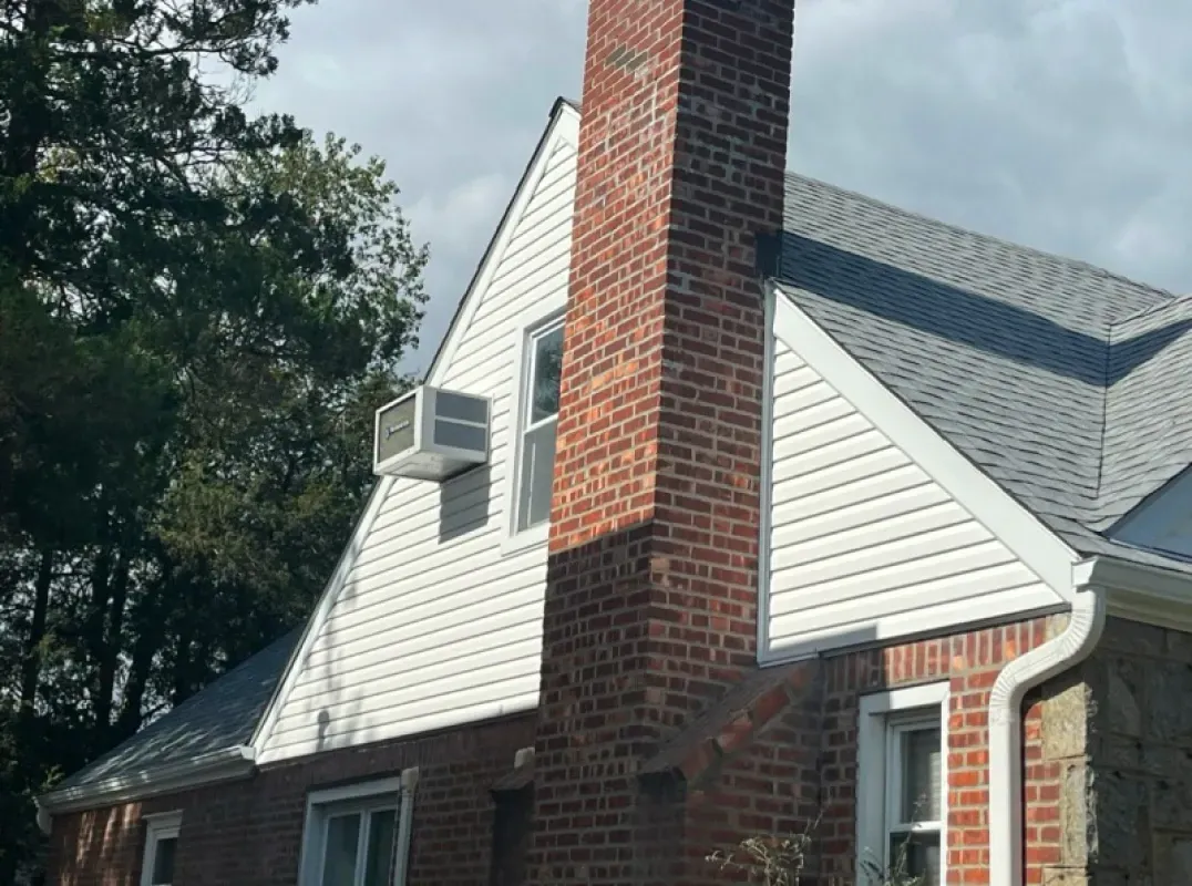 A brick chimney rises along the gable end of a house with white vinyl siding and a window air conditioning unit.