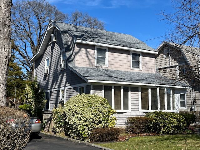 A two-story grey shingled house with a full-width enclosed front porch, green shrubs, and a driveway on a sunny day.