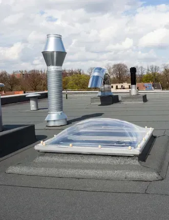 A flat roof with a skylight and several metal ventilation pipes against a cloudy sky.