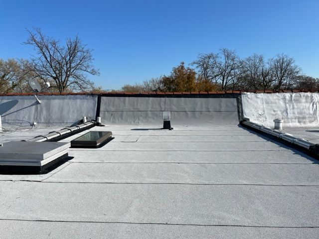 A flat gray roof under a clear blue sky, featuring a skylight and rooftop vents, with trees visible in the background.