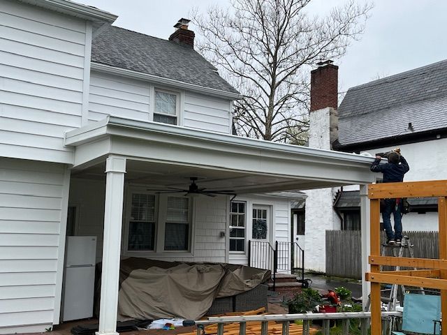A person on a ladder installs gutters on a covered patio attached to a two-story white house.
