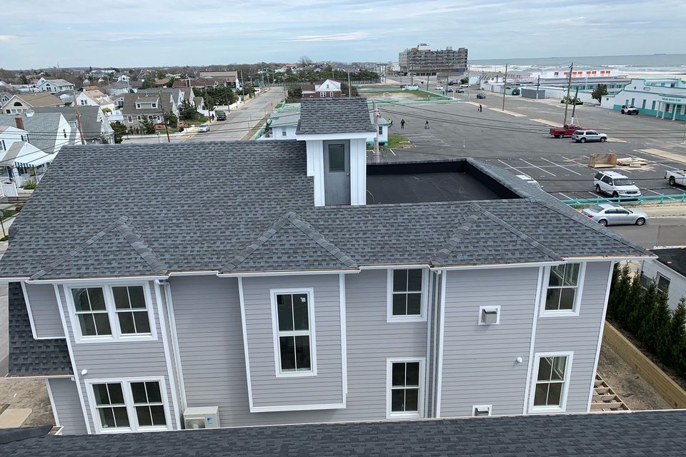 A high-angle view of a gray, two-story house with a shingled roof, set in a coastal neighborhood near a parking lot.