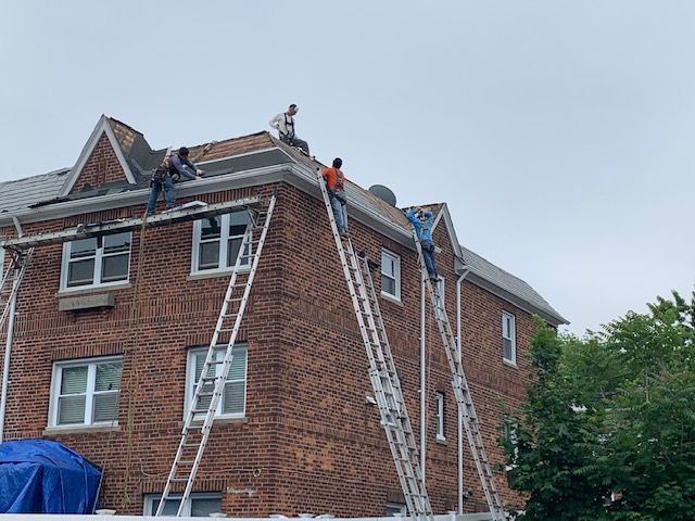 Four workers on ladders and the roof of a multi-story brick building, performing roof repairs.