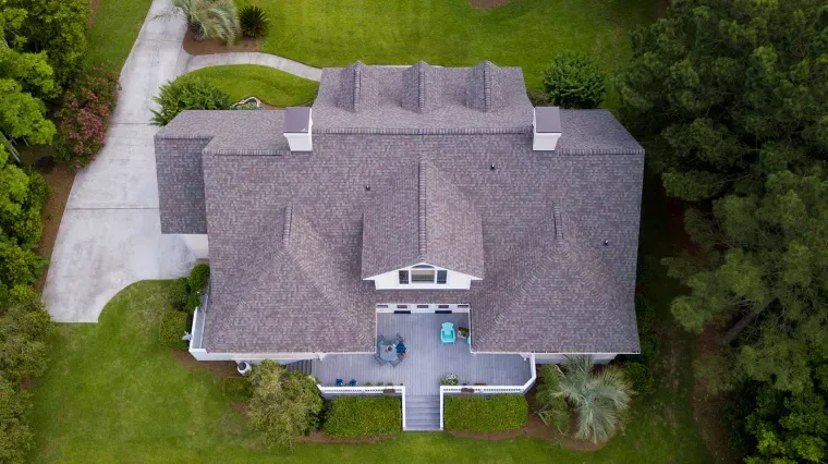 Aerial view of a house with a shingled roof, a back patio, and a driveway, surrounded by green lawns and trees.
