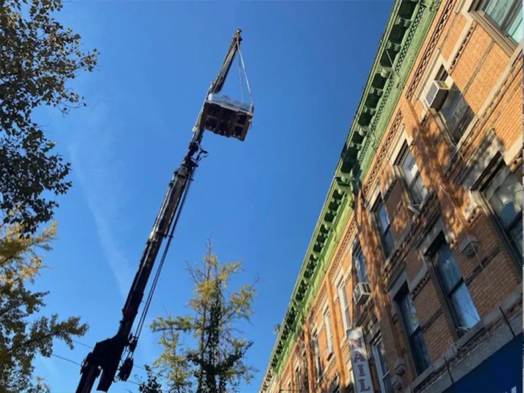 A construction crane lifts a work platform toward the roofline of an old brick building against a bright blue sky.