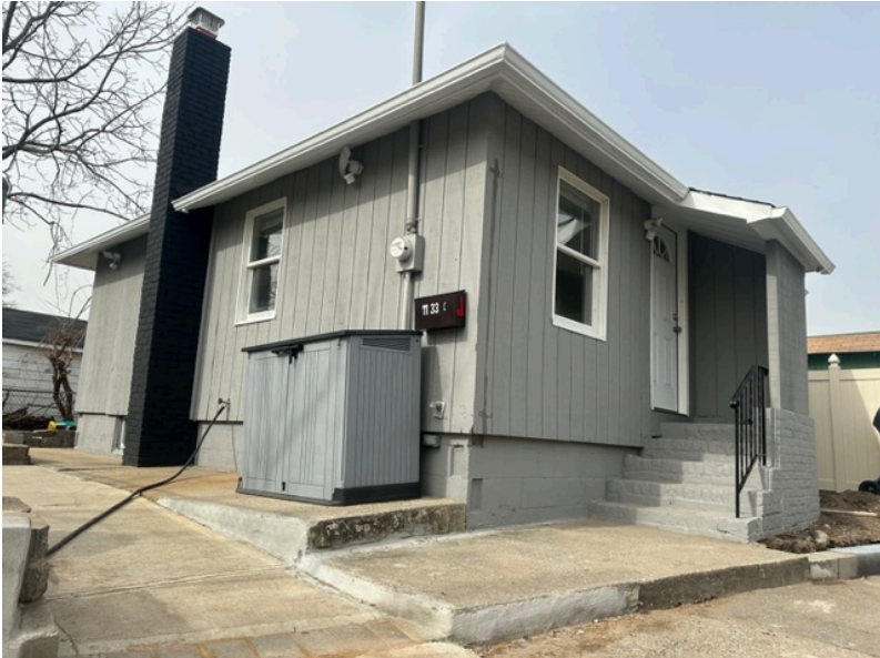 A small, gray, single-story house with a tall black chimney, concrete stairs, and a grey storage shed in the yard.