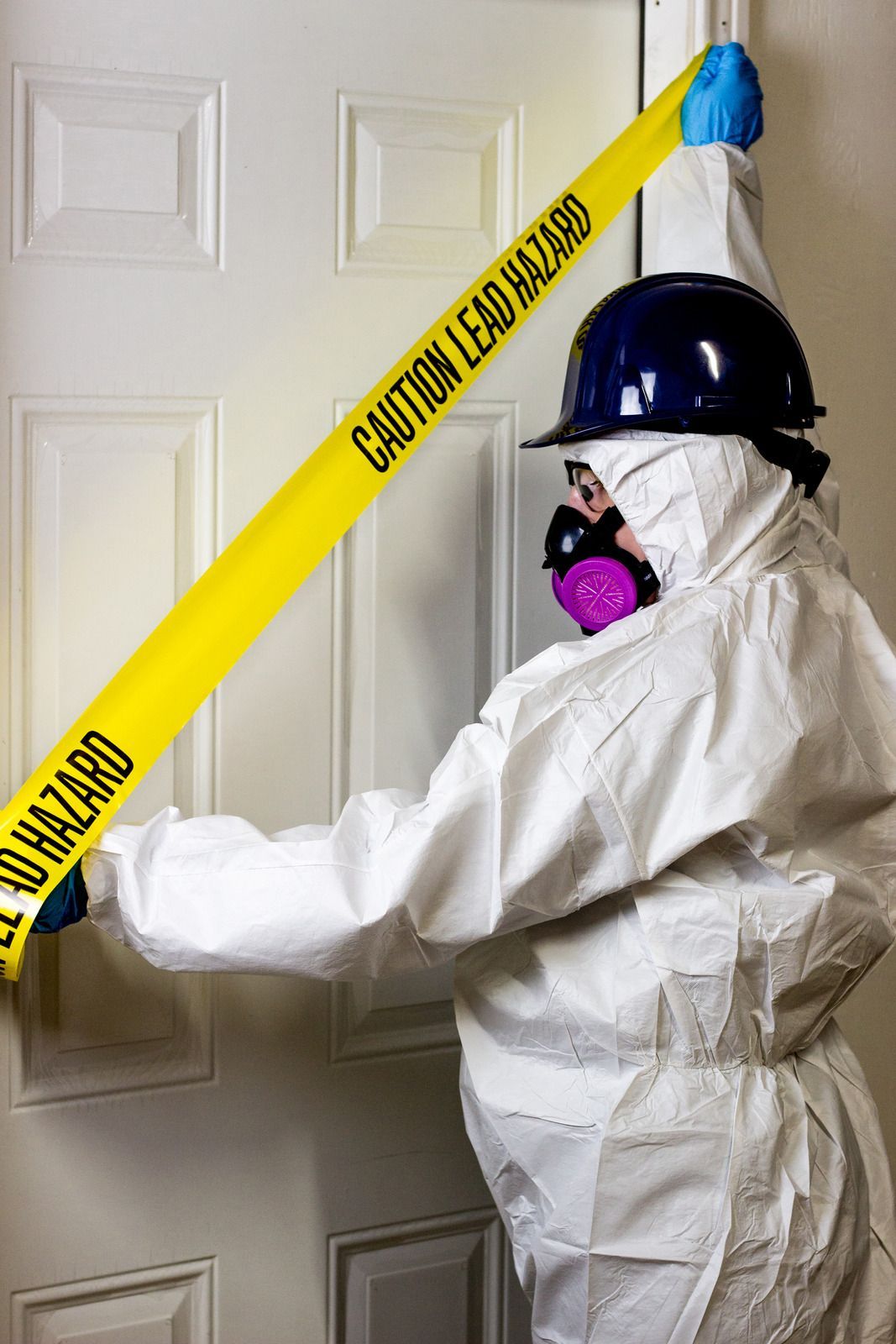 A person in a protective suit is holding a yellow tape that says caution lead hazard.