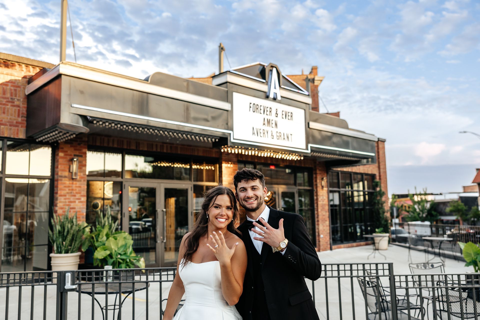 Couple in wedding attire showing off rings in front of a brick building.