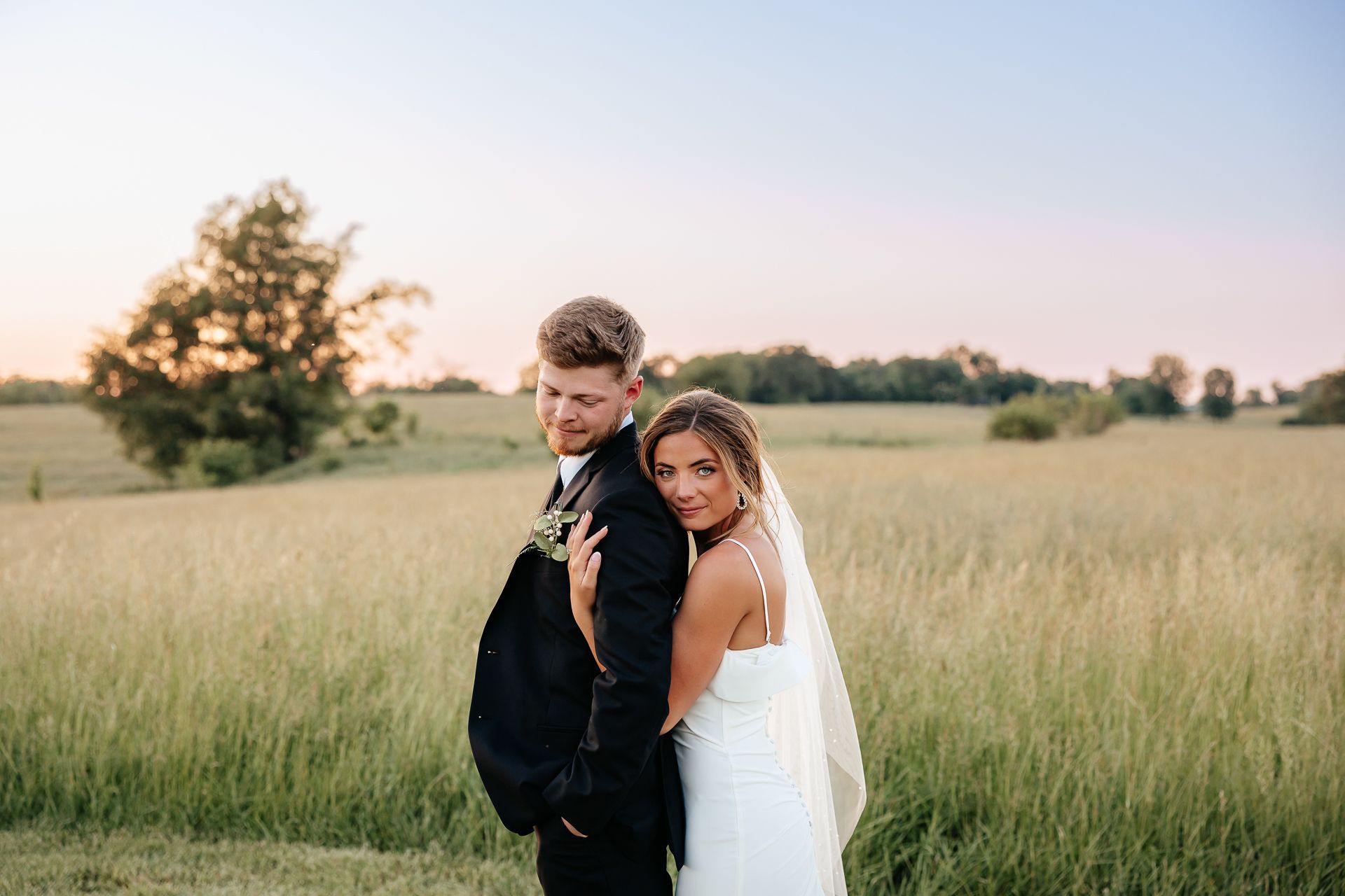 Bride and groom embrace in a field at sunset. The bride's head rests on the groom's shoulder.