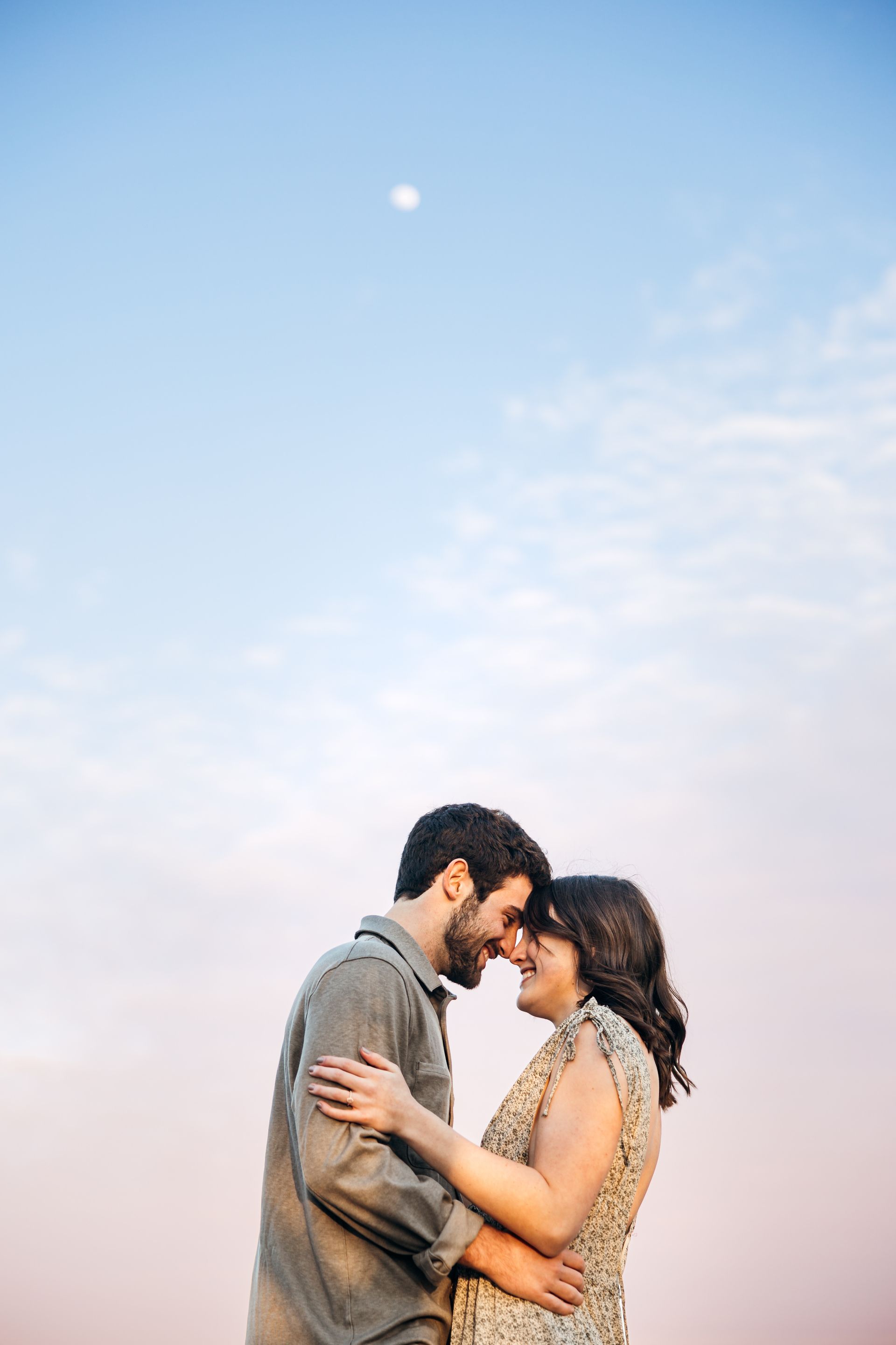 Couple embracing, foreheads touching, smiling against a sky with a faint moon and pink/blue hues.