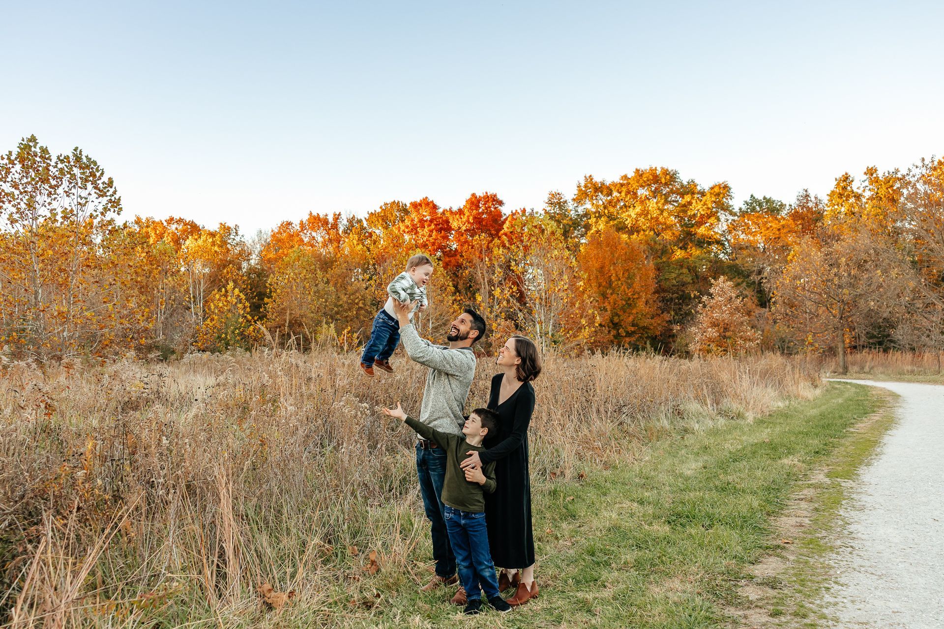 Family of four playing outdoors, dad tossing baby in air, autumn foliage in background.