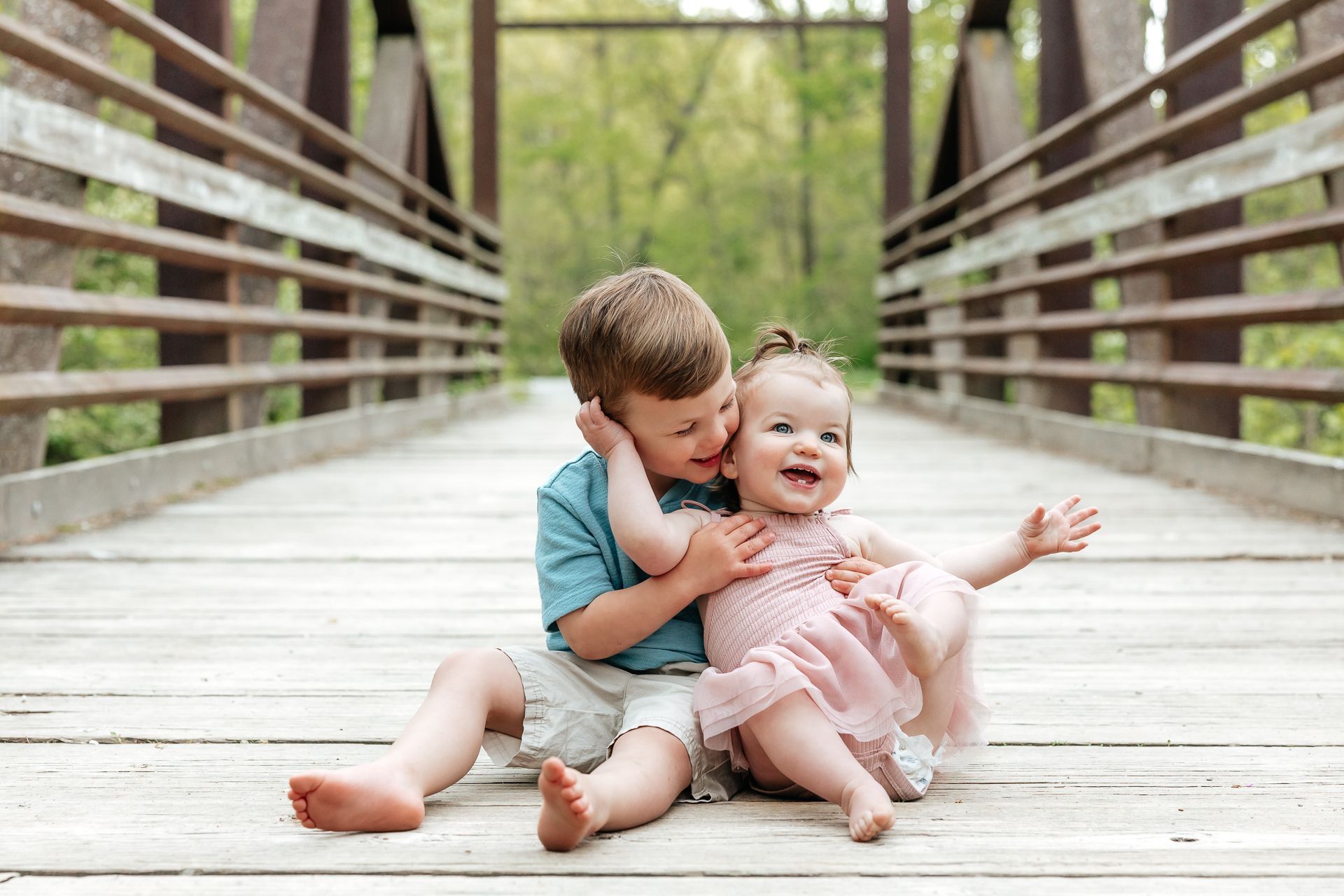 Two children smiling on a wooden bridge, one hugging the other. Green trees in background.