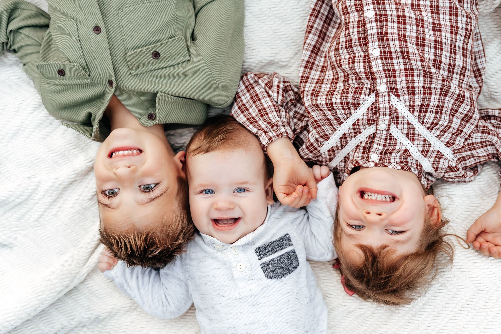 Three children lying on their backs, smiling at the camera. One is a baby, and the others are older.