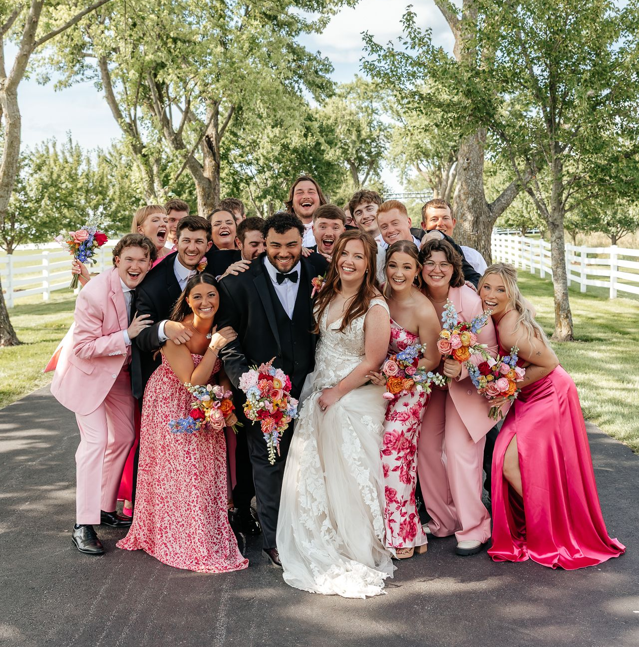 Wedding party portrait, bride and groom surrounded by smiling friends outdoors, holding bouquets.