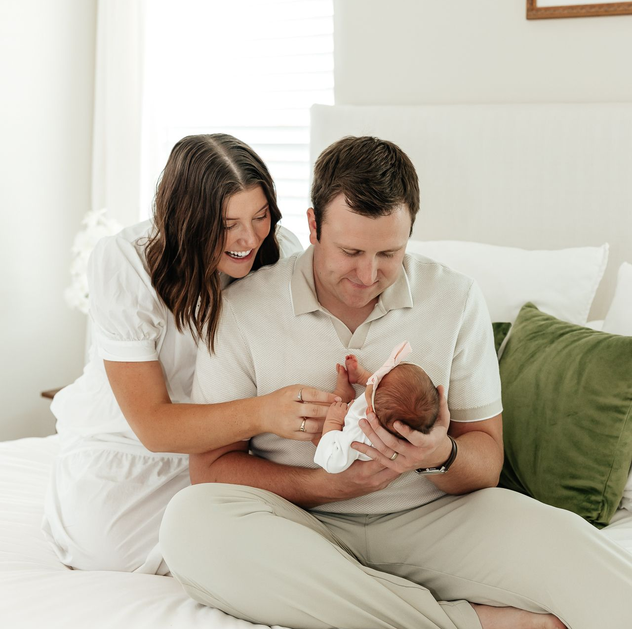Parents holding newborn baby on a bed; mother smiles, father looks down, pillows.