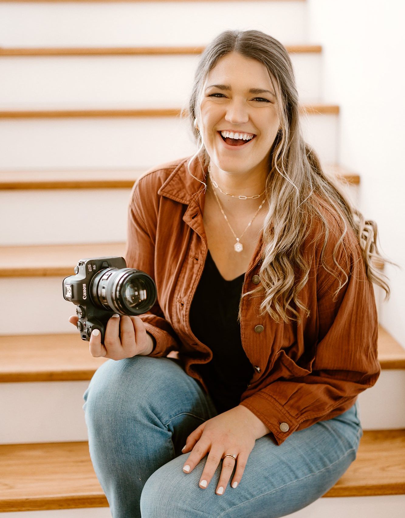 Woman smiling, holding a camera, sitting on stairs, wearing a brown jacket and jeans.