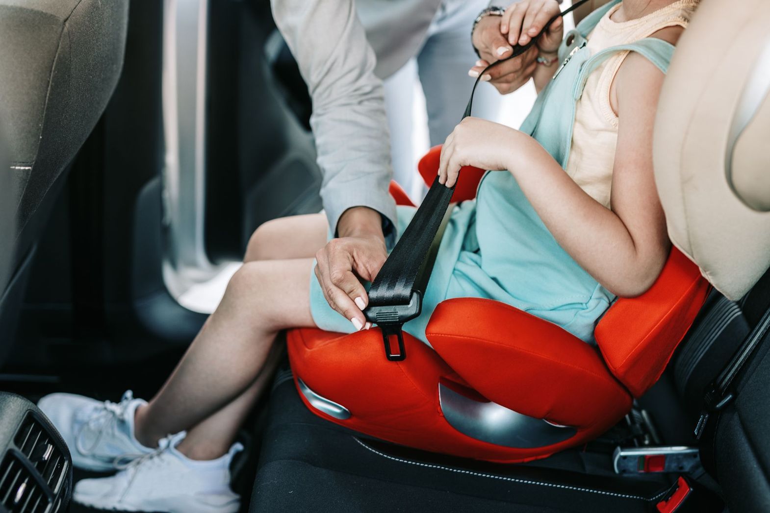 Child buckled into a red car seat while an adult fastens the harness in a vehicle Child buckled into a red car seat while an adult fastens the harness in a vehicle