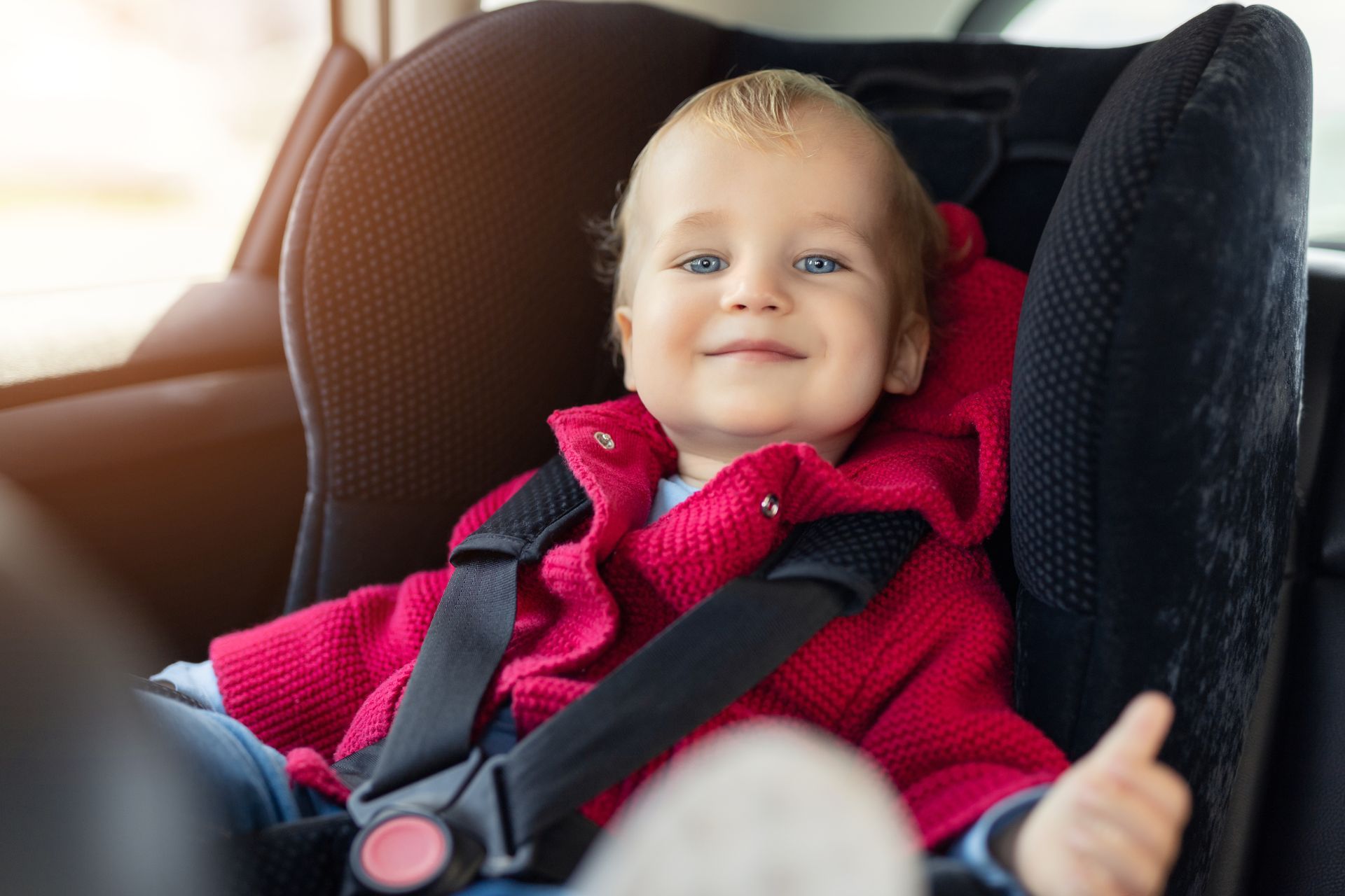 Smiling child strapped into a car seat, wearing a bright pink sweater and black harness. Smiling child strapped into a car seat, wearing a bright pink sweater and black harness.