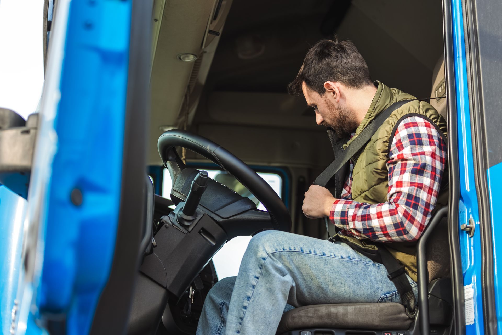 Man seated in a blue truck cab, adjusting his seat belt or harness, with plaid shirt and jeans. Man seated in a blue truck cab, adjusting his seat belt or harness, with plaid shirt and jeans.