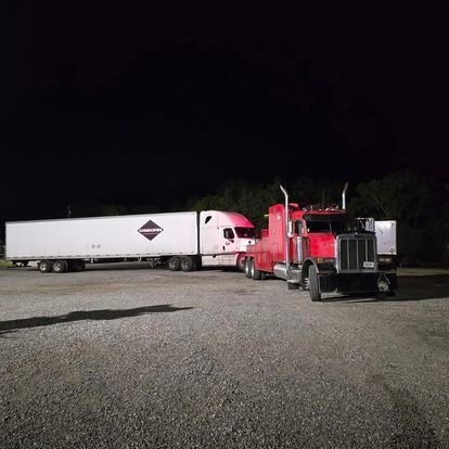 Two semi trucks are parked in a gravel lot at night.
