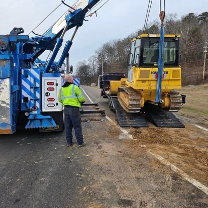 A man is standing next to a tow truck that is carrying a bulldozer.