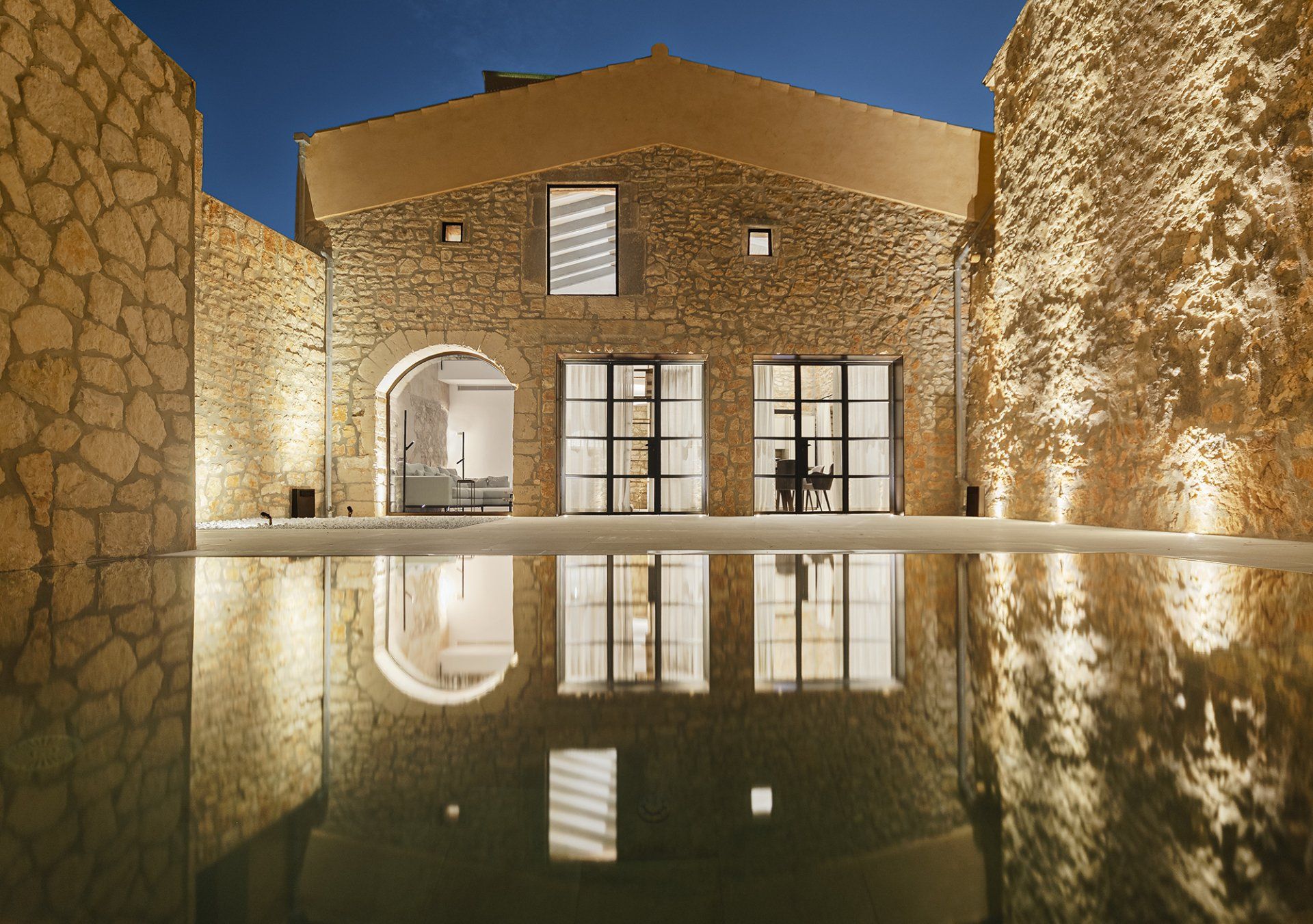 Casa de piedra con patio y luces cálidas que se reflejan en un estanque tranquilo al atardecer.