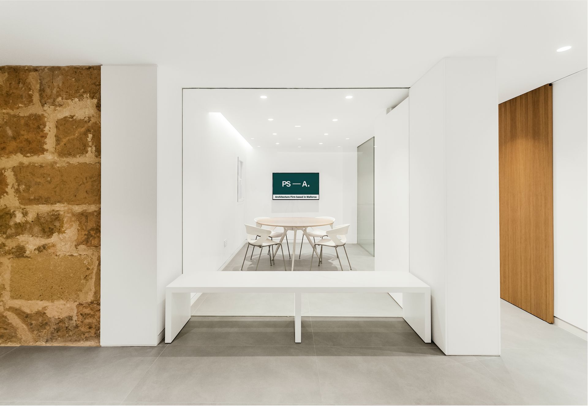 Comedor luminoso y minimalista con mesa y sillas blancas, pared de piedra y detalles en la puerta de madera.