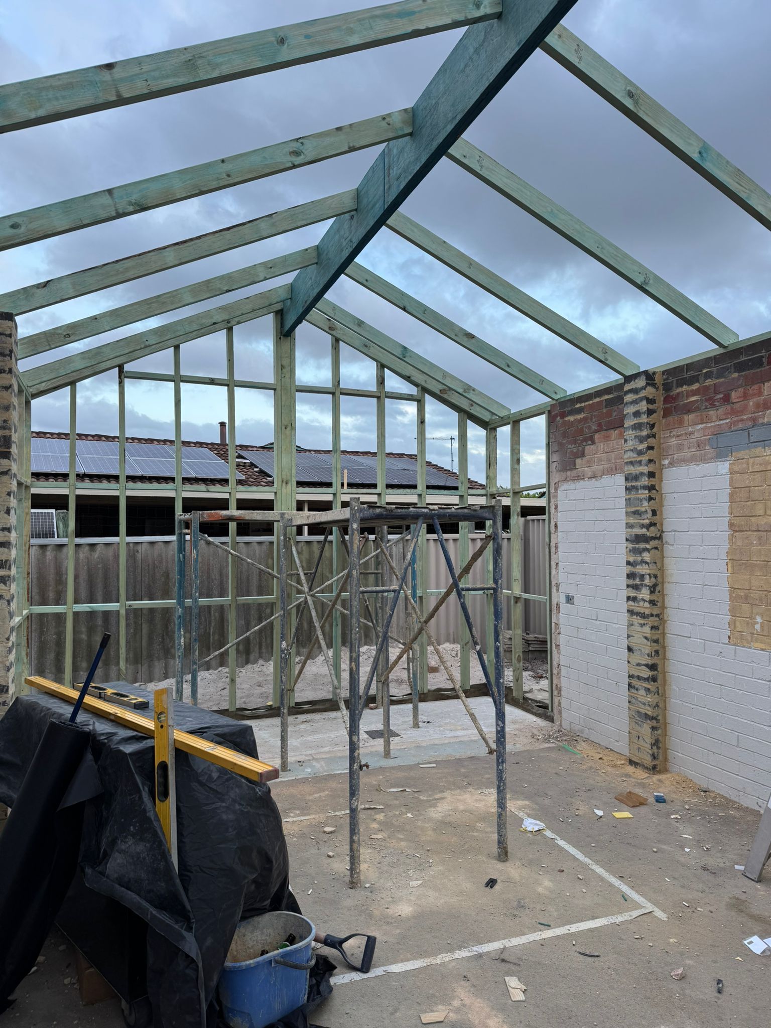 Partially built steel roof frame in a backyard construction site, with scaffolding and brick walls nearby