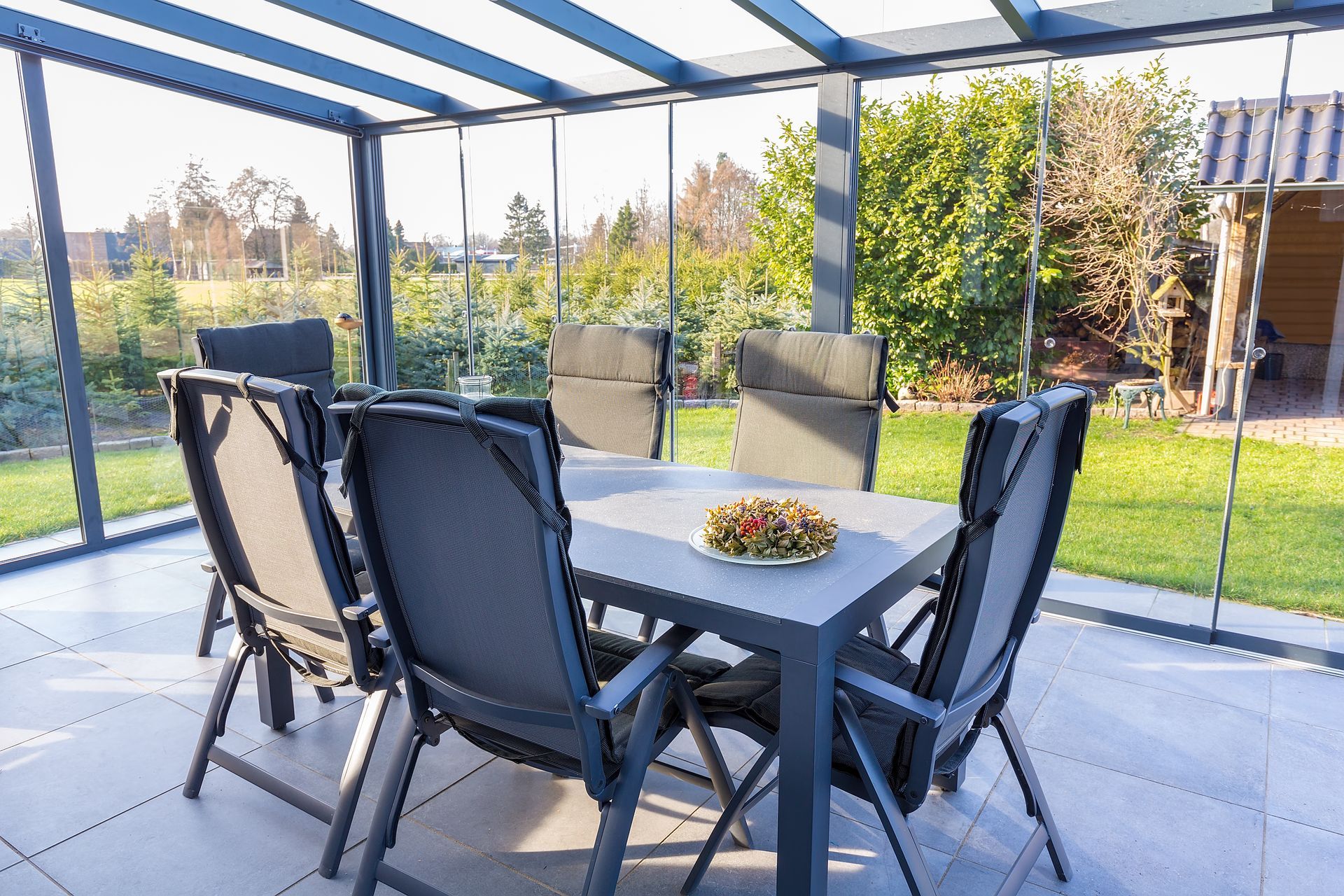Covered patio dining table with six black chairs and glass wind screens overlooking a garden