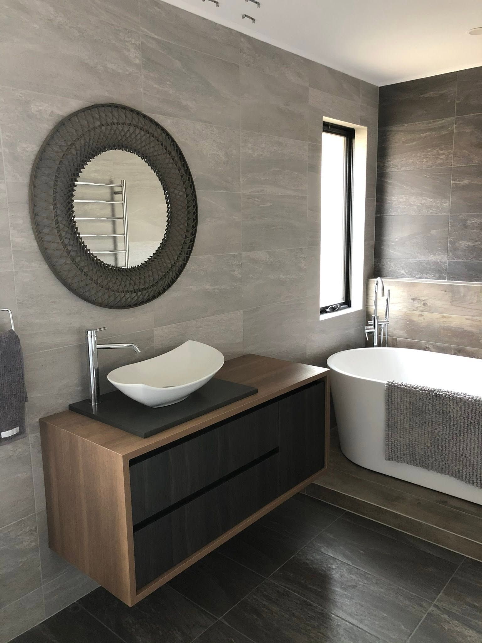 Modern bathroom with gray tile, wooden vanity, oval mirror, vessel sink, and a bathtub by the window