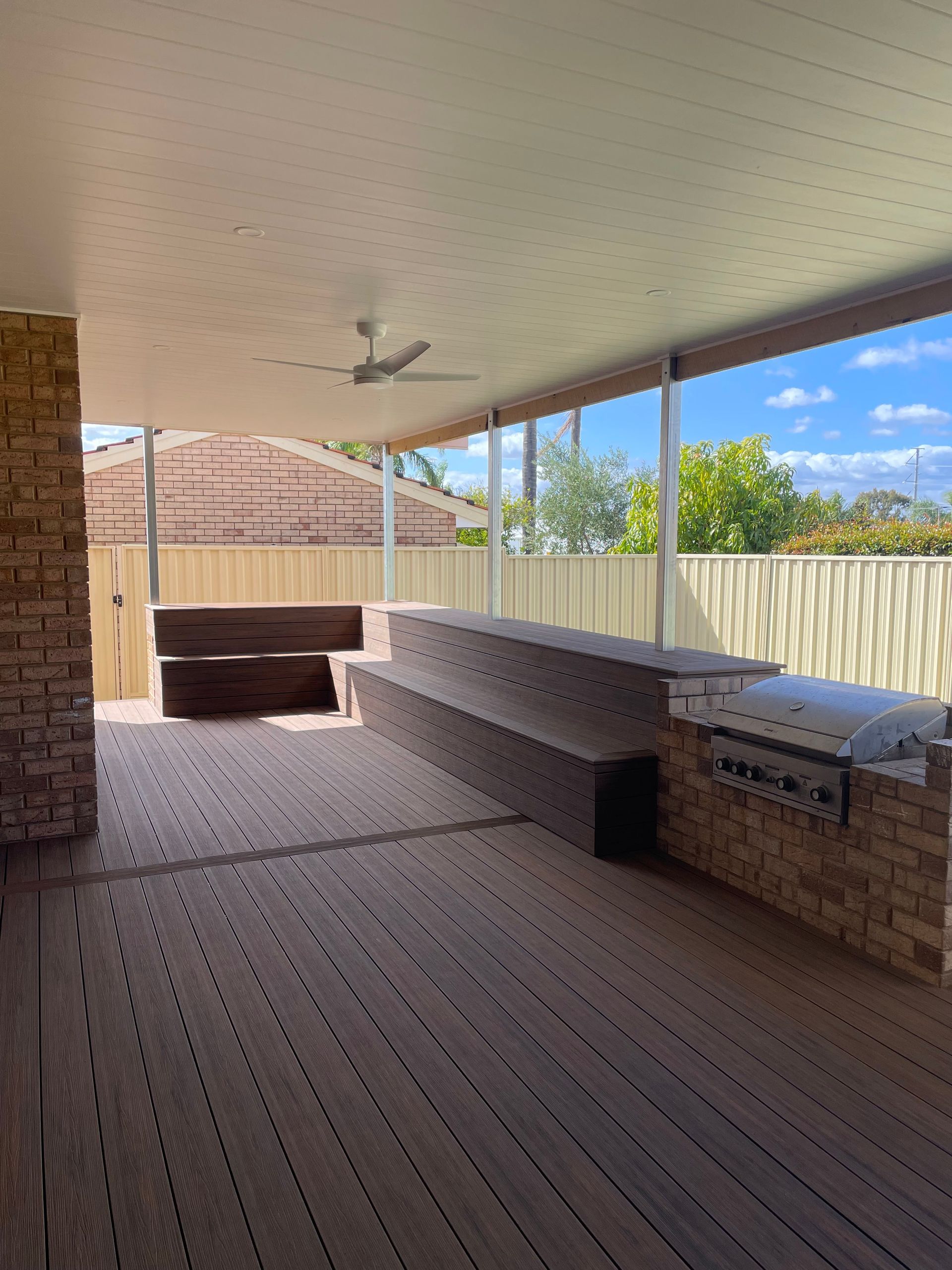Covered patio with brick seating, brown deck flooring, ceiling fan, and view of a fenced yard