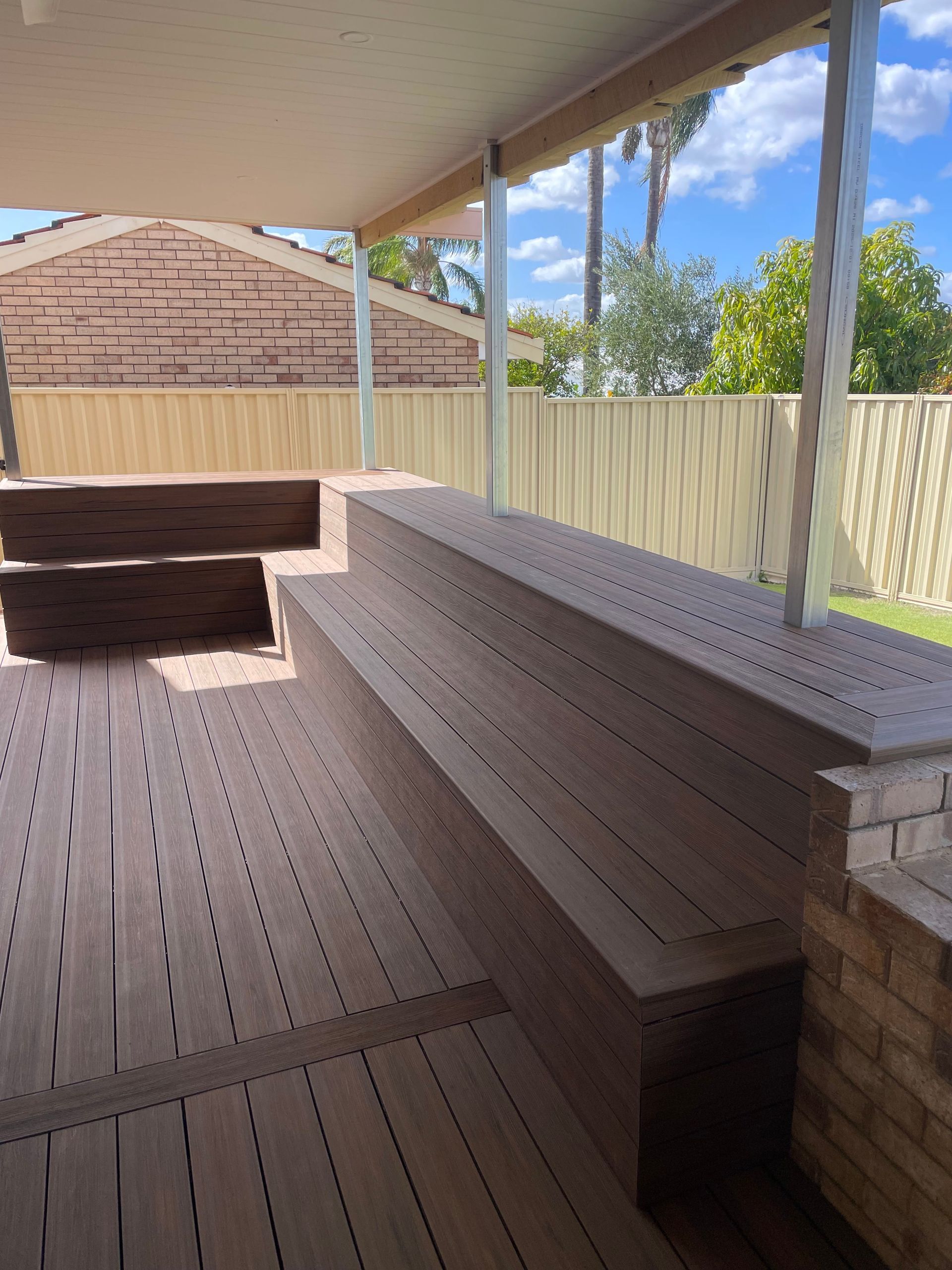 Covered patio with built-in wooden bench seating and a brick planter wall overlooking a fenced yard.