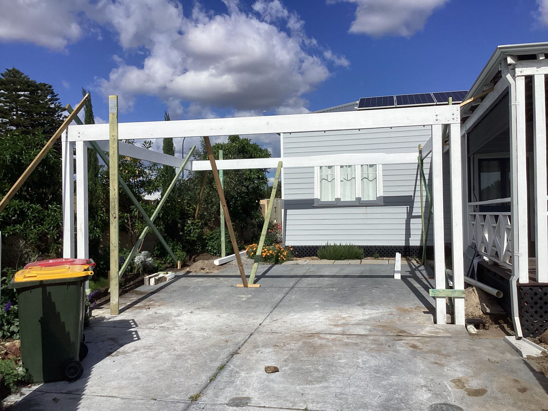 Backyard patio with white pergola frame, concrete slab, green bins, and a shed under a blue sky.