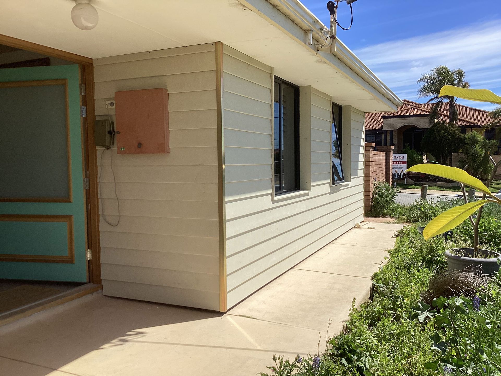 Single-story house exterior with beige siding, blue door, and a concrete walkway beside a garden.