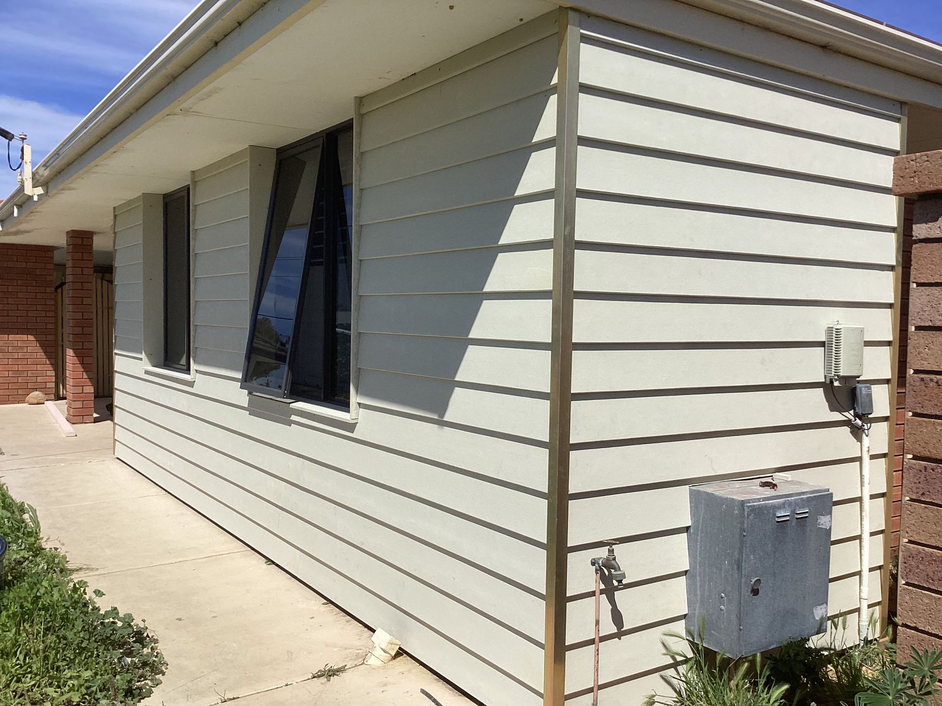 Side view of a beige house with windows, concrete walkway, and utility boxes along the exterior wall