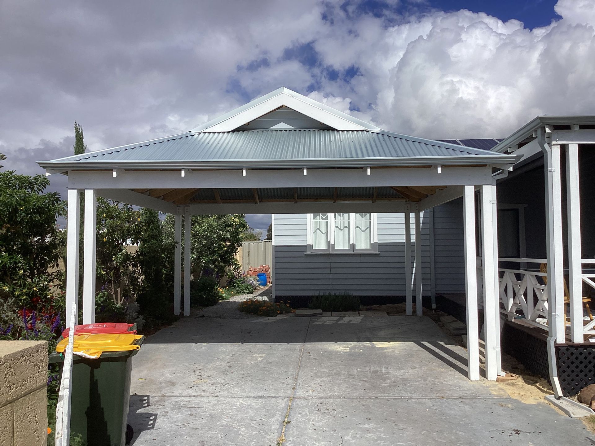 Carport driveway beside a house under a cloudy sky, with trash bins at left.