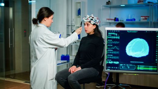 Clinician adjusting EEG cap on seated patient in a lab, with brain scan displayed on monitor.
