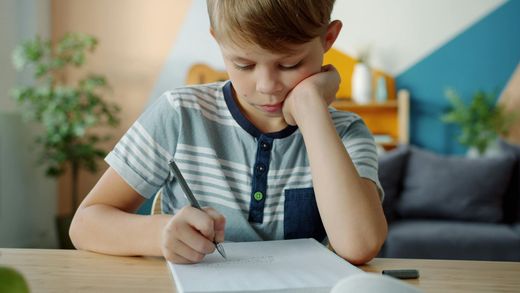 Child writing at a table, resting head on hand, in a bright living room.
