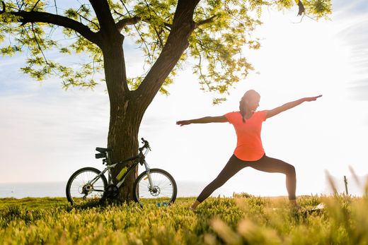 Person doing a yoga pose beside a tree with a bicycle in a sunny field
