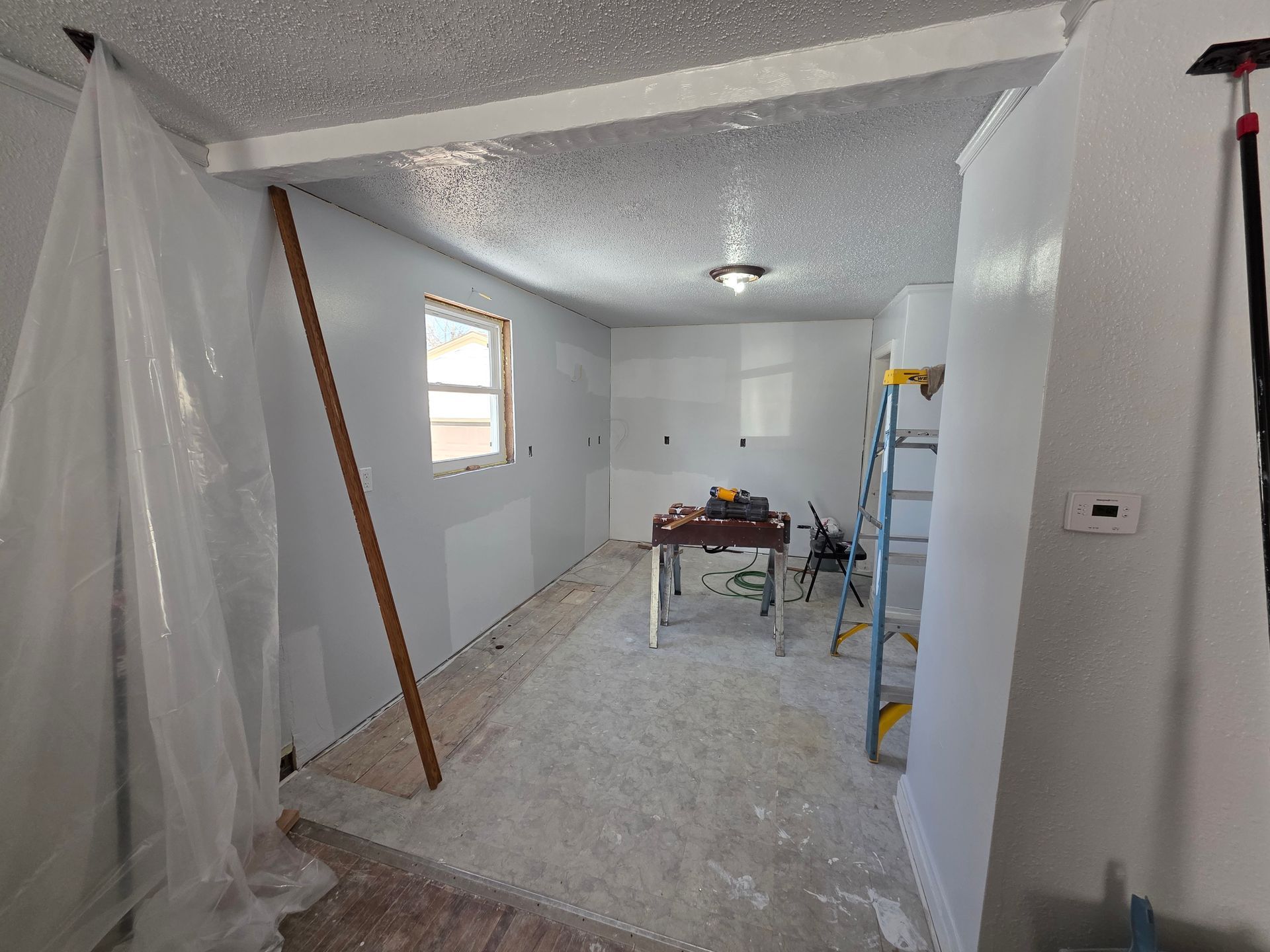 Partially renovated room with plastic sheeting, white walls, a window, and work tools on a table