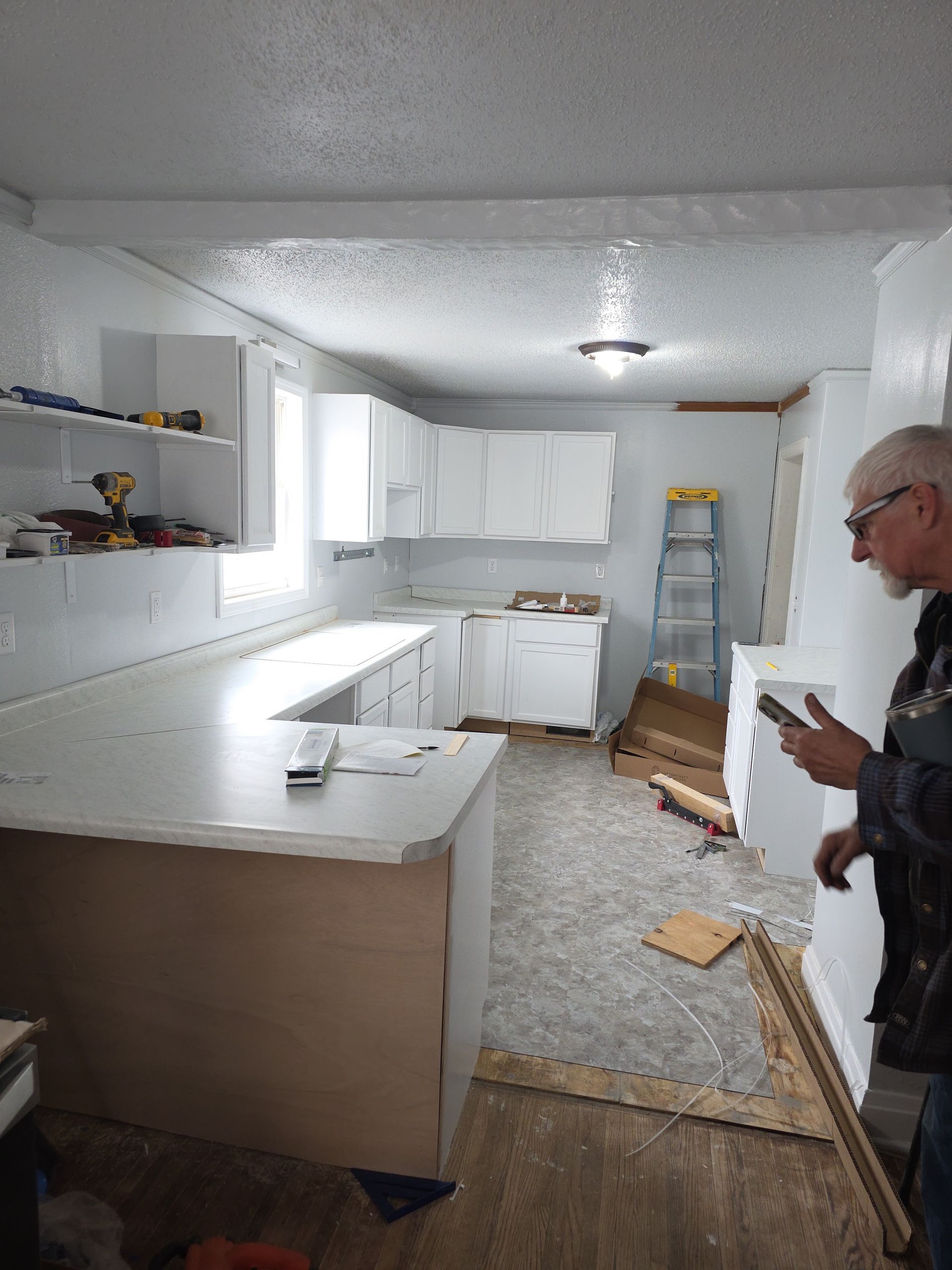 Renovating kitchen with white cabinets, ladder, and a man standing on the right holding a tool.