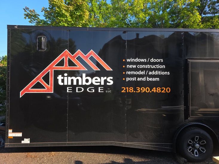 Black Timbers Edge trailer with orange logo, service list, and phone number parked outdoors.