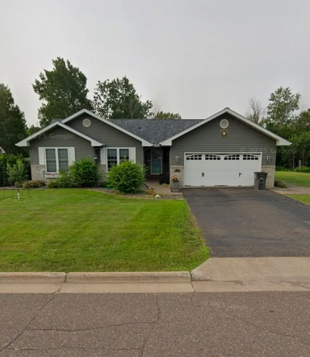 Single-story suburban house with gray siding, a two-car garage, and a green lawn under an overcast sky.