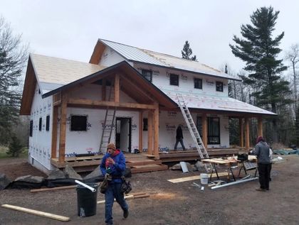 Partially built house with workers, ladders, and construction materials in a wooded yard