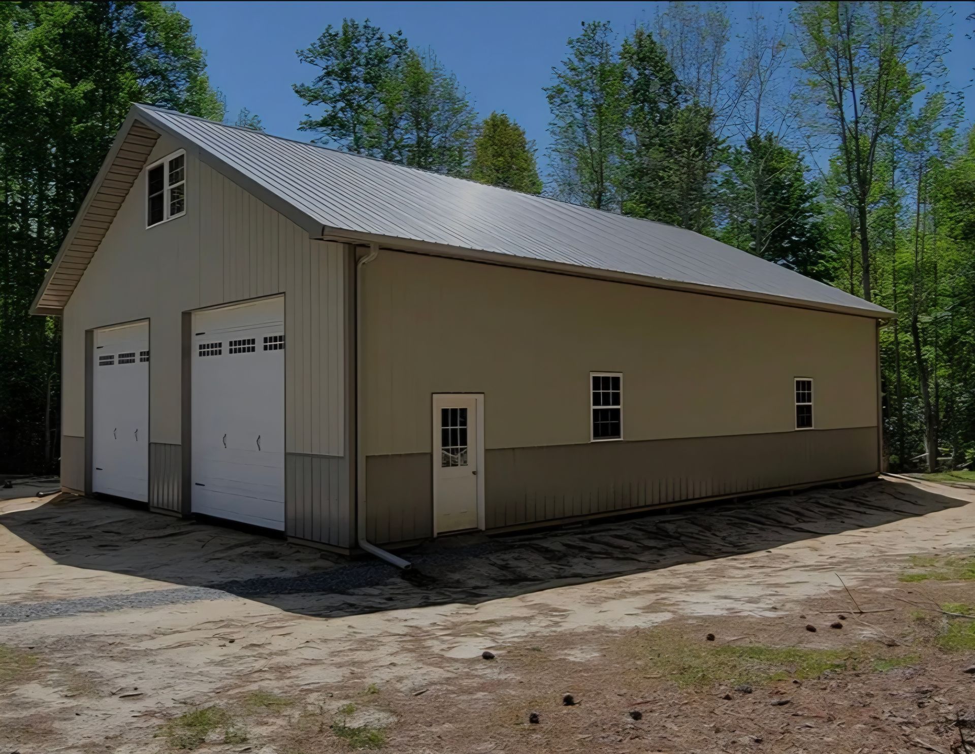 Tan garage with white double doors, side windows, and a metal roof in a wooded clearing