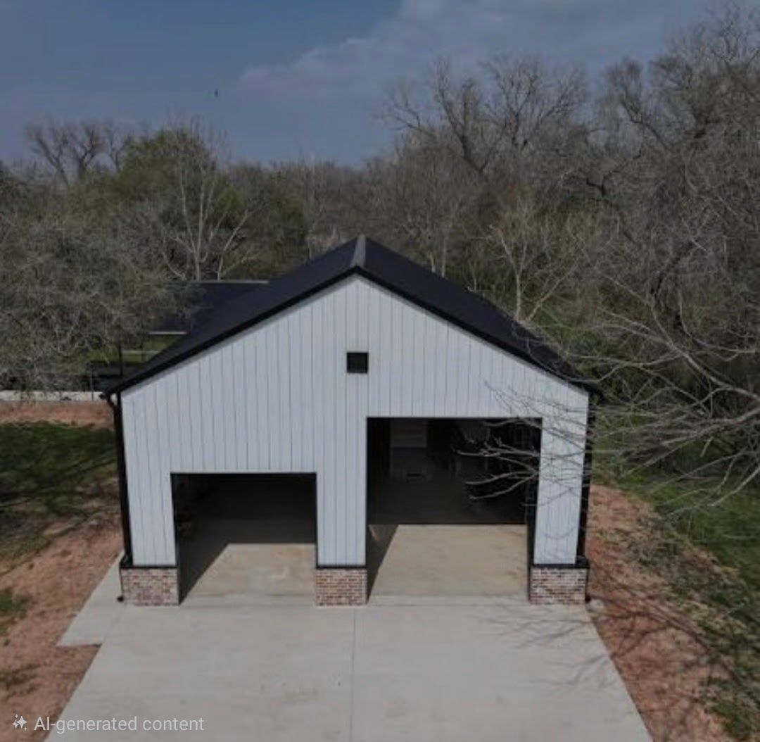 Detached white barn with black roof and two open garage bays beside a concrete driveway, surrounded by trees