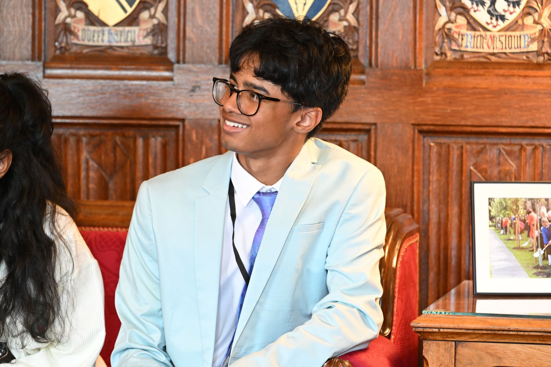 Person in a light blue blazer smiling while seated in a wood-paneled room with red chairs.